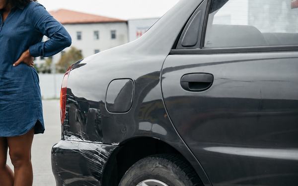 A woman standing beside her car, looking at a dented rear panel.
