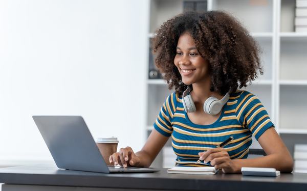 A young woman working at a laptop with a notebook and coffee beside her.