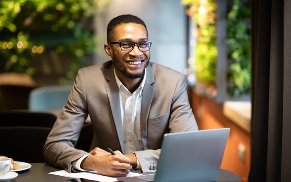A man smiling as he works on a laptop at a café table.